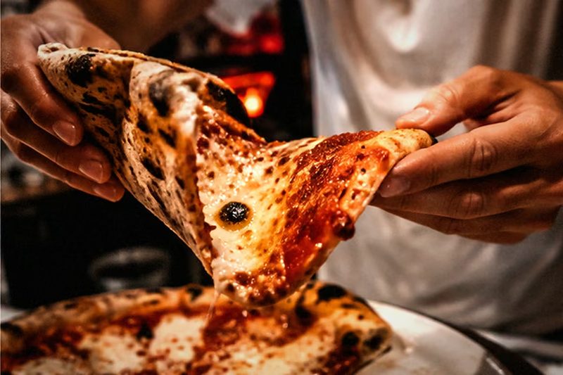 Zwei Hände heben ein Stück Pizza an, der Käse zieht Fäden