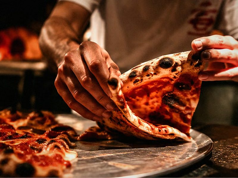 Hände am Tresen heben ein Stück Pizza vom Blech, im Hintergrund der Ofen