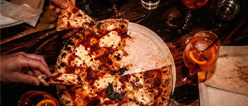 Tisch von oben bei Nacht: angeschnittene Pizza, Gläser mit orangefarbenen Drinks, Kerzenlicht